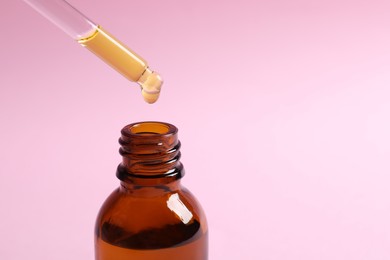 Dripping cosmetic oil from pipette into bottle on pink background, closeup. Space for text