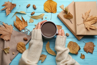 Woman with cup of hot drink at light blue wooden table, top view. Cozy autumn atmosphere