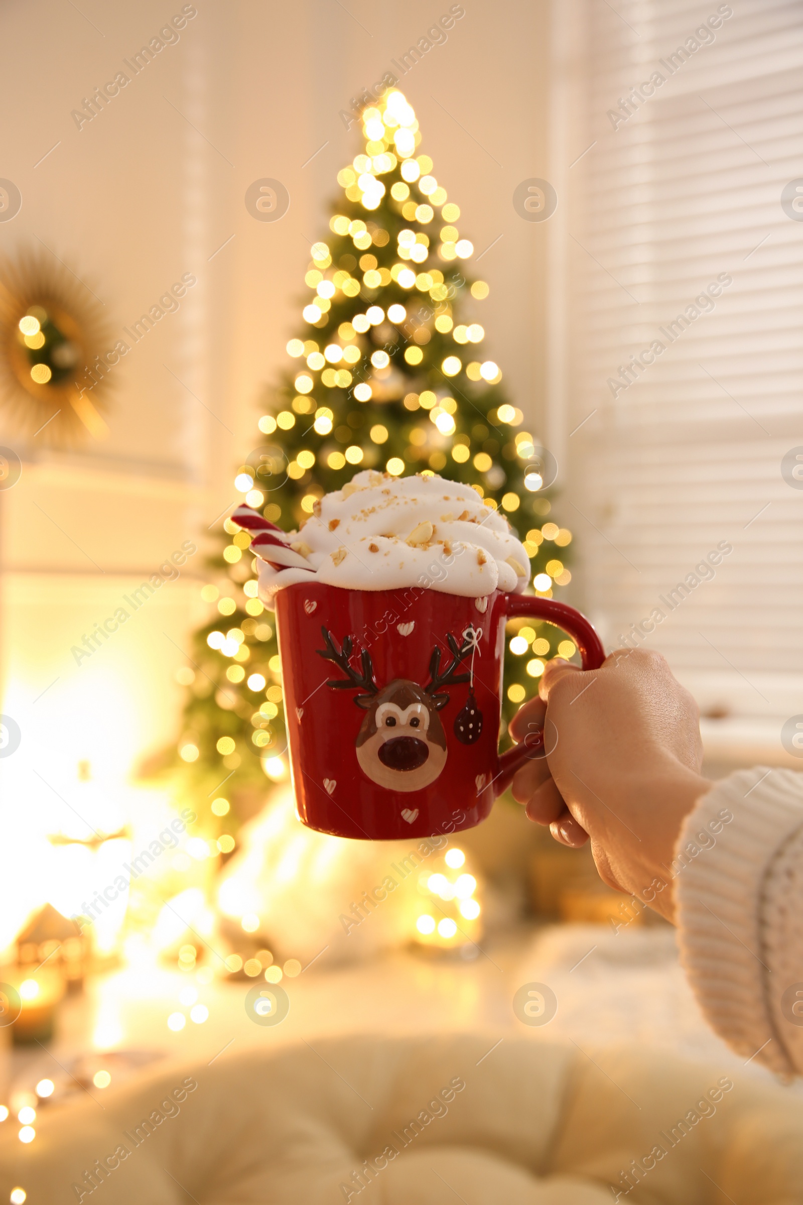 Photo of Woman holding cup of delicious drink with whipped cream near Christmas tree indoors, closeup