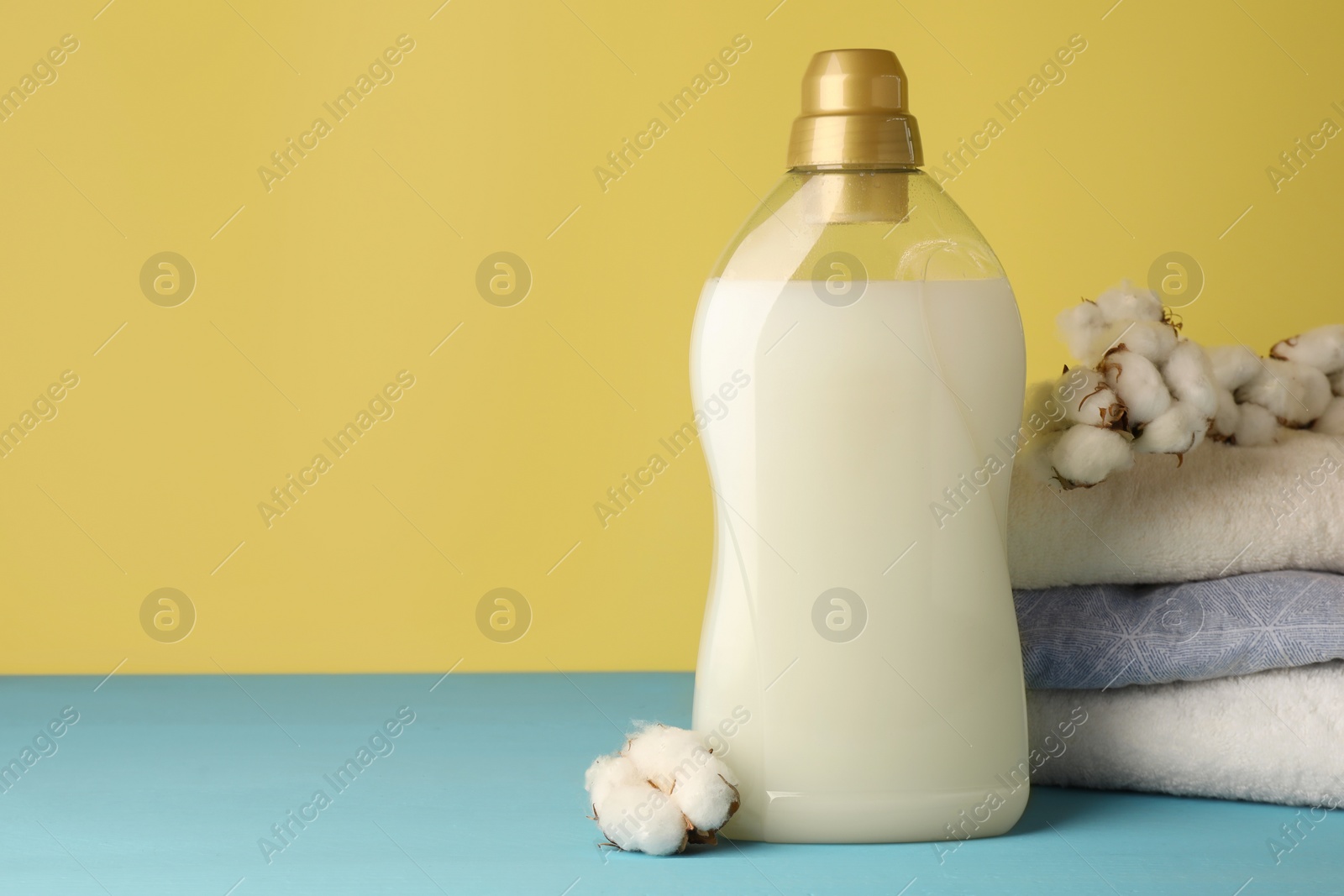 Photo of Bottle of fabric softener, towels and cotton flowers on light blue table, space for text