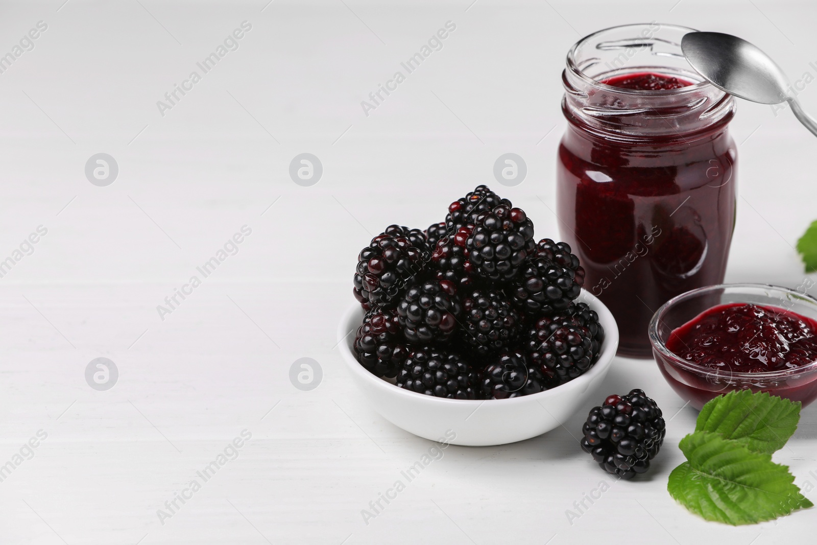 Photo of Fresh ripe blackberries, tasty jam and leaves on white wooden table. Space for text