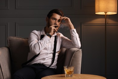 Photo of Tired man smoking cigar and resting at home in evening. Glass of whiskey on table near him
