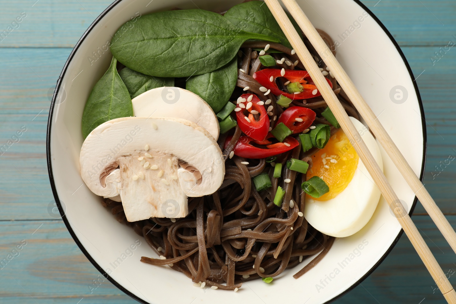 Photo of Tasty buckwheat noodles (soba) with chili pepper, egg, mushrooms and chopsticks on light blue wooden table, top view