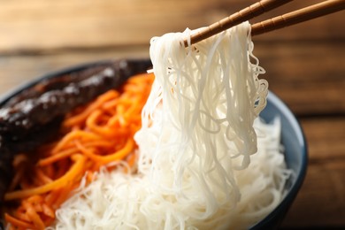 Chopsticks with tasty cooked rice noodles over bowl, closeup