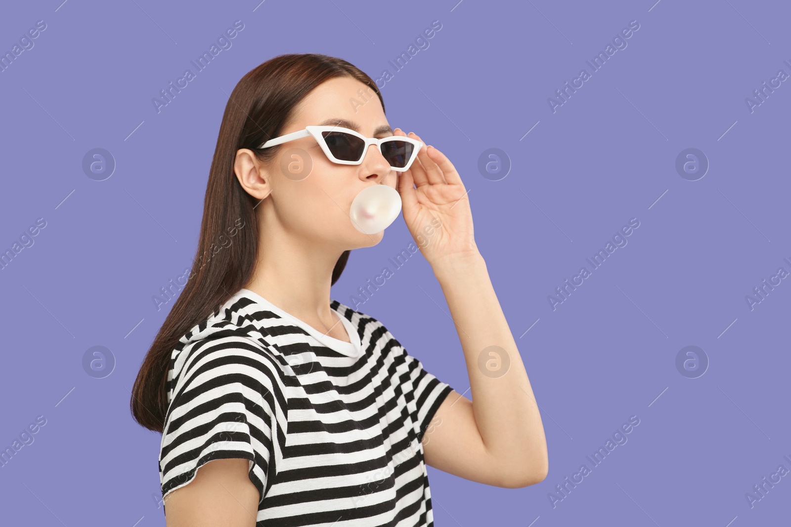 Photo of Beautiful woman in sunglasses blowing bubble gum on light purple background