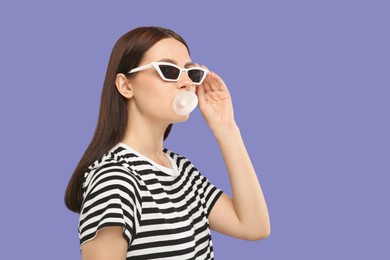 Photo of Beautiful woman in sunglasses blowing bubble gum on light purple background