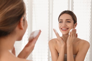 Young woman applying cleansing foam onto her face near mirror in bathroom