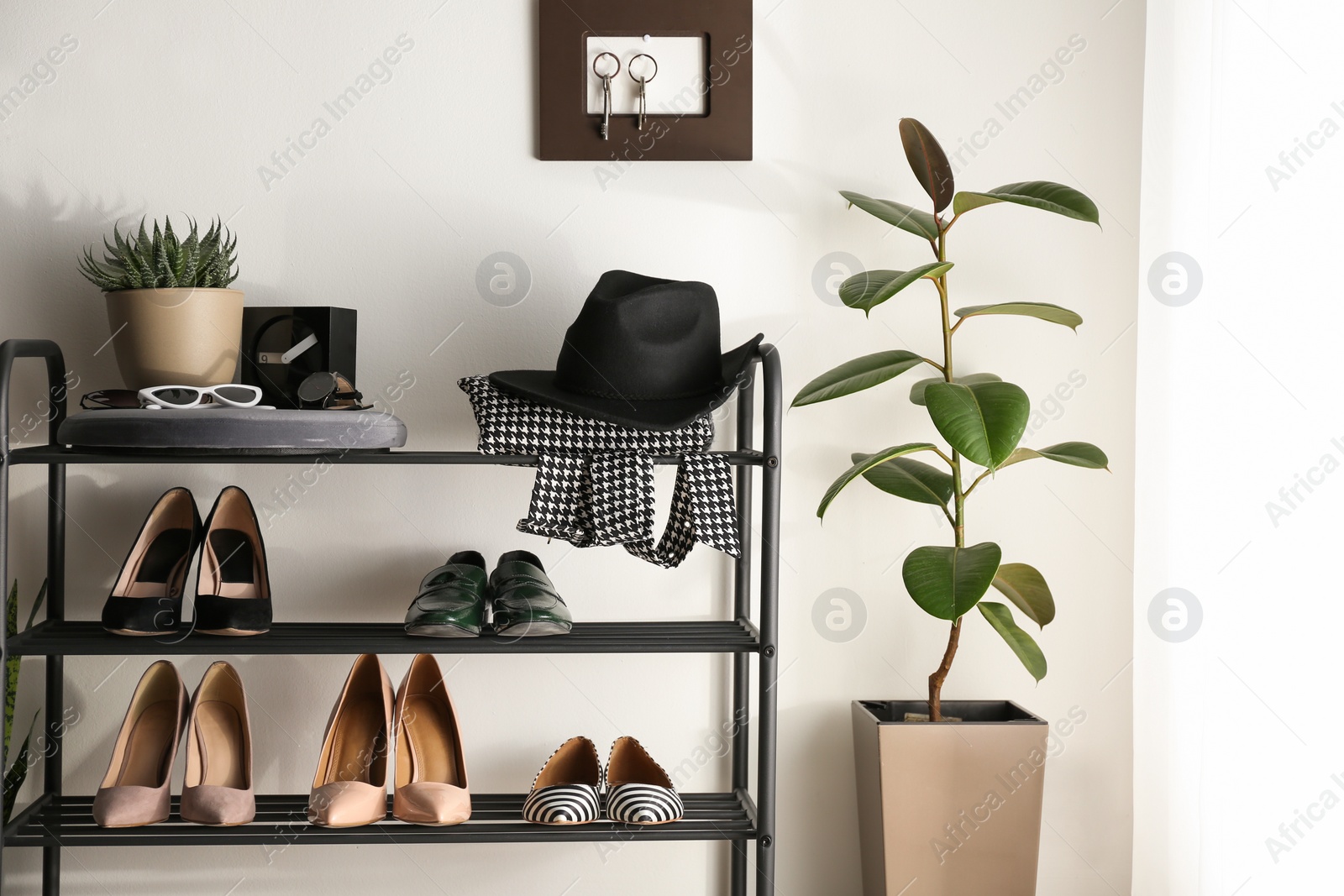 Photo of Black shelving unit with shoes and different accessories near white wall in hall