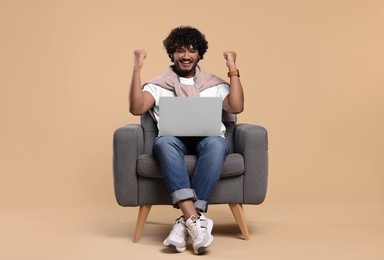 Happy man with laptop sitting in armchair on beige background