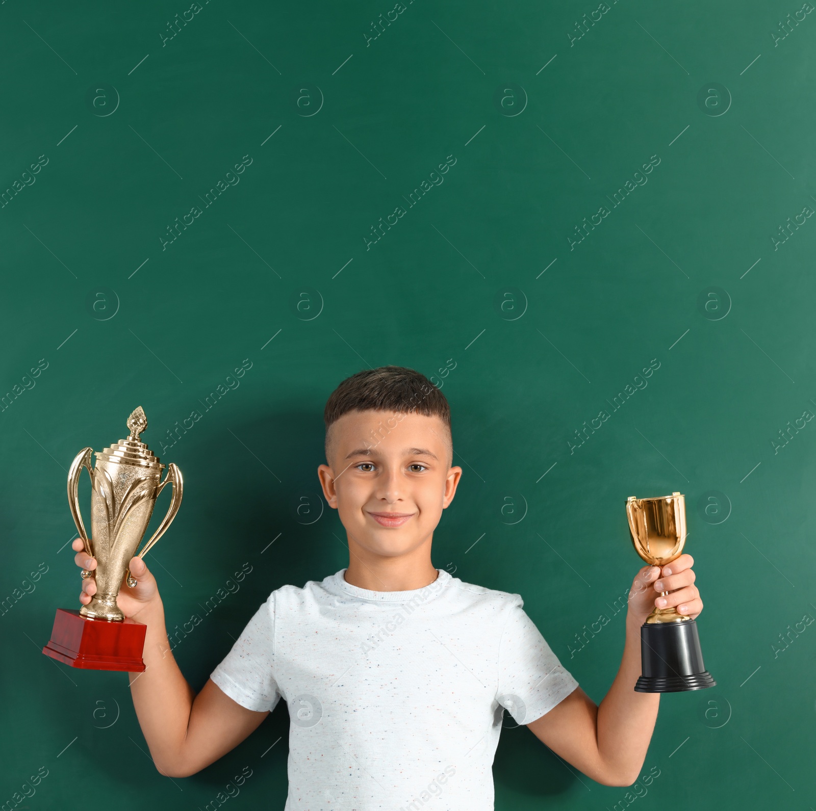 Photo of Happy boy with golden winning cups near chalkboard