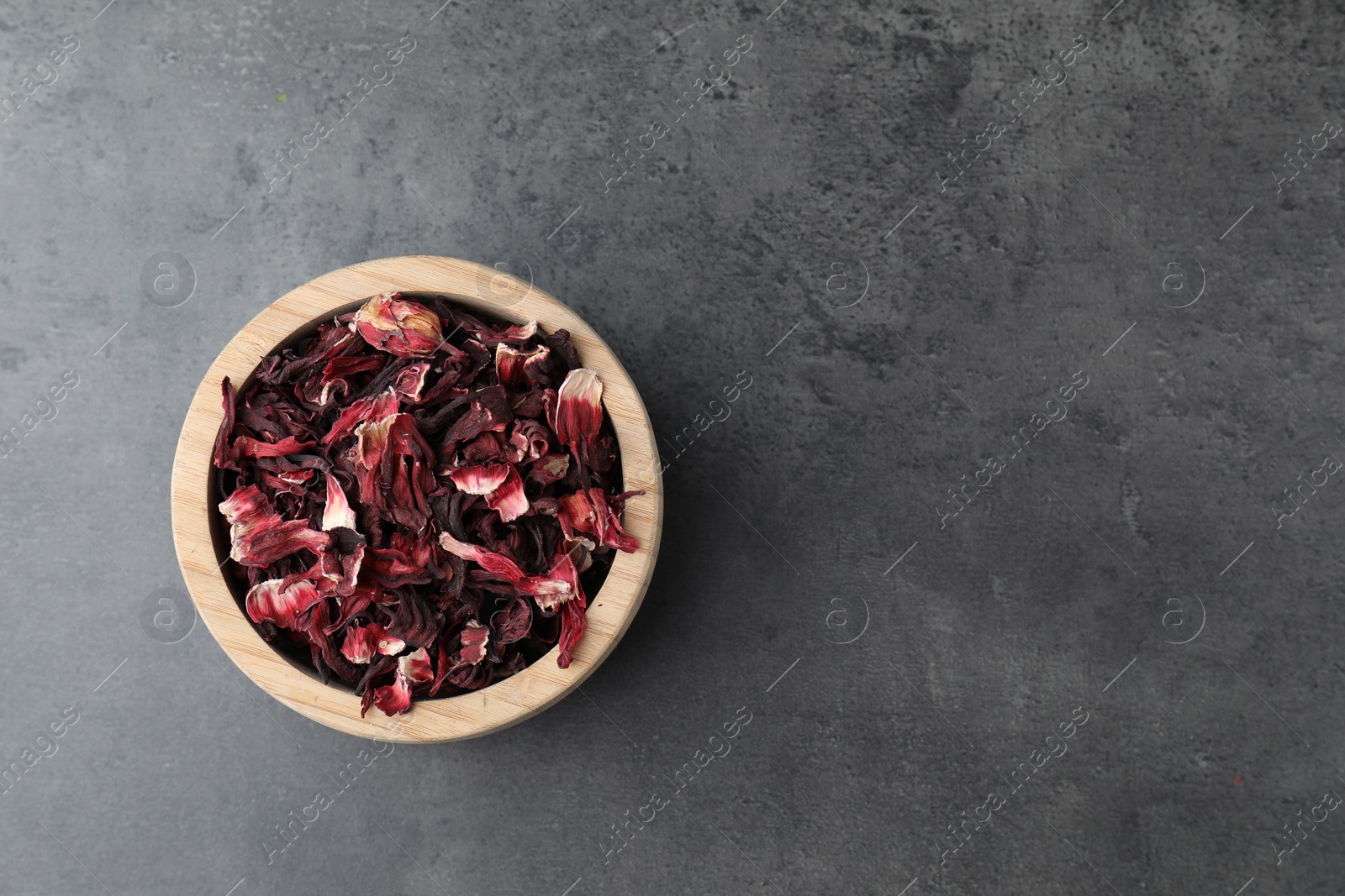 Photo of Hibiscus tea. Wooden bowl with dried roselle calyces on grey table, top view. Space for text