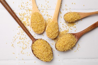 Photo of Spoons with raw bulgur on white tiled table, flat lay