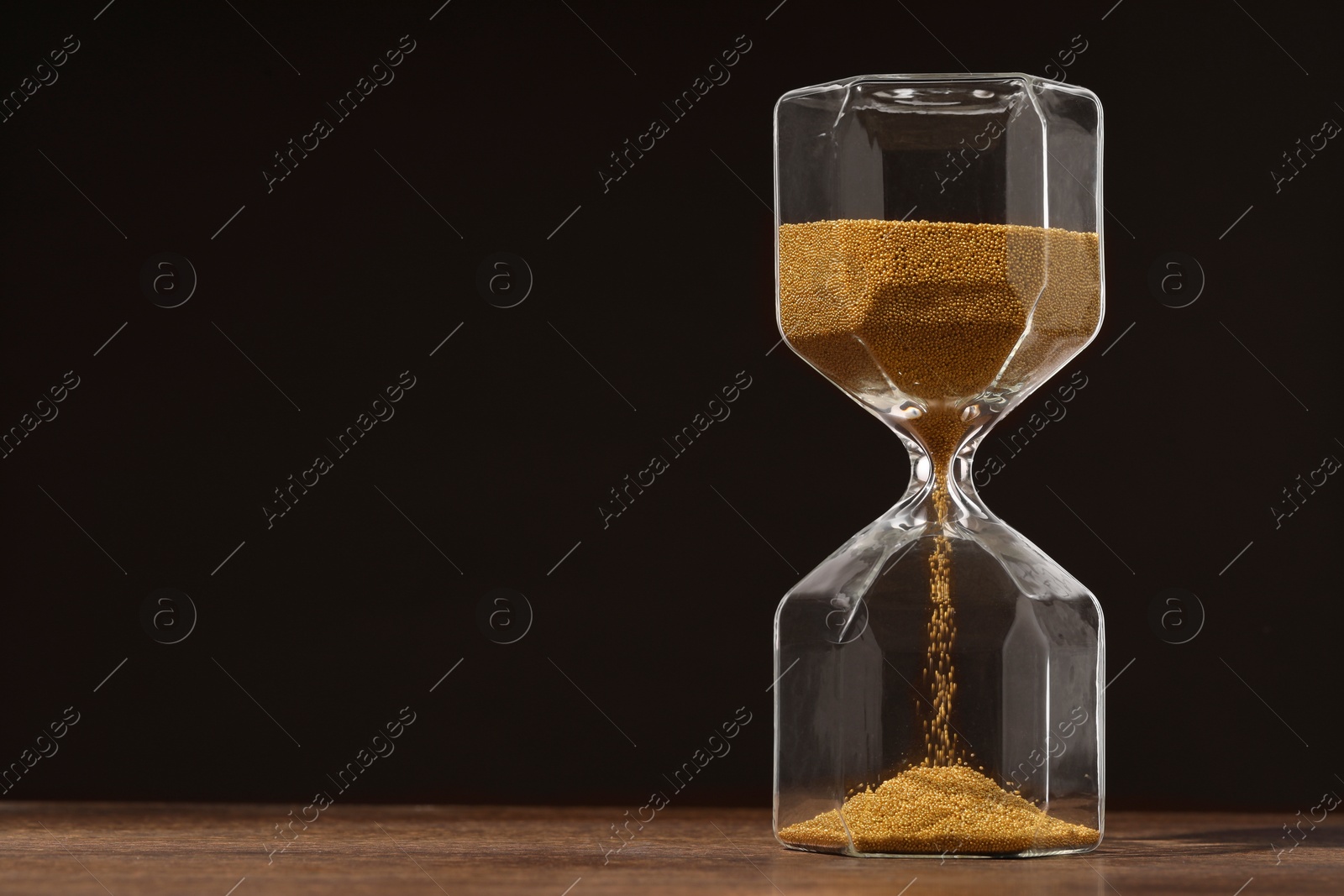 Photo of Hourglass with flowing sand on wooden table against dark brown background, space for text