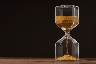 Hourglass with flowing sand on wooden table against dark brown background, space for text