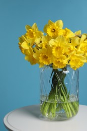 Photo of Beautiful daffodils in vase on white table against light blue background