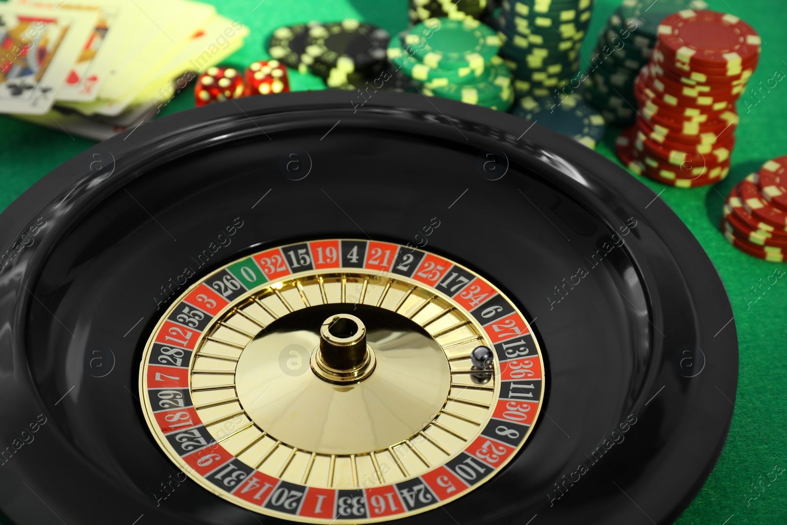 Photo of Roulette wheel with ball, playing cards and chips on green table, closeup. Casino game