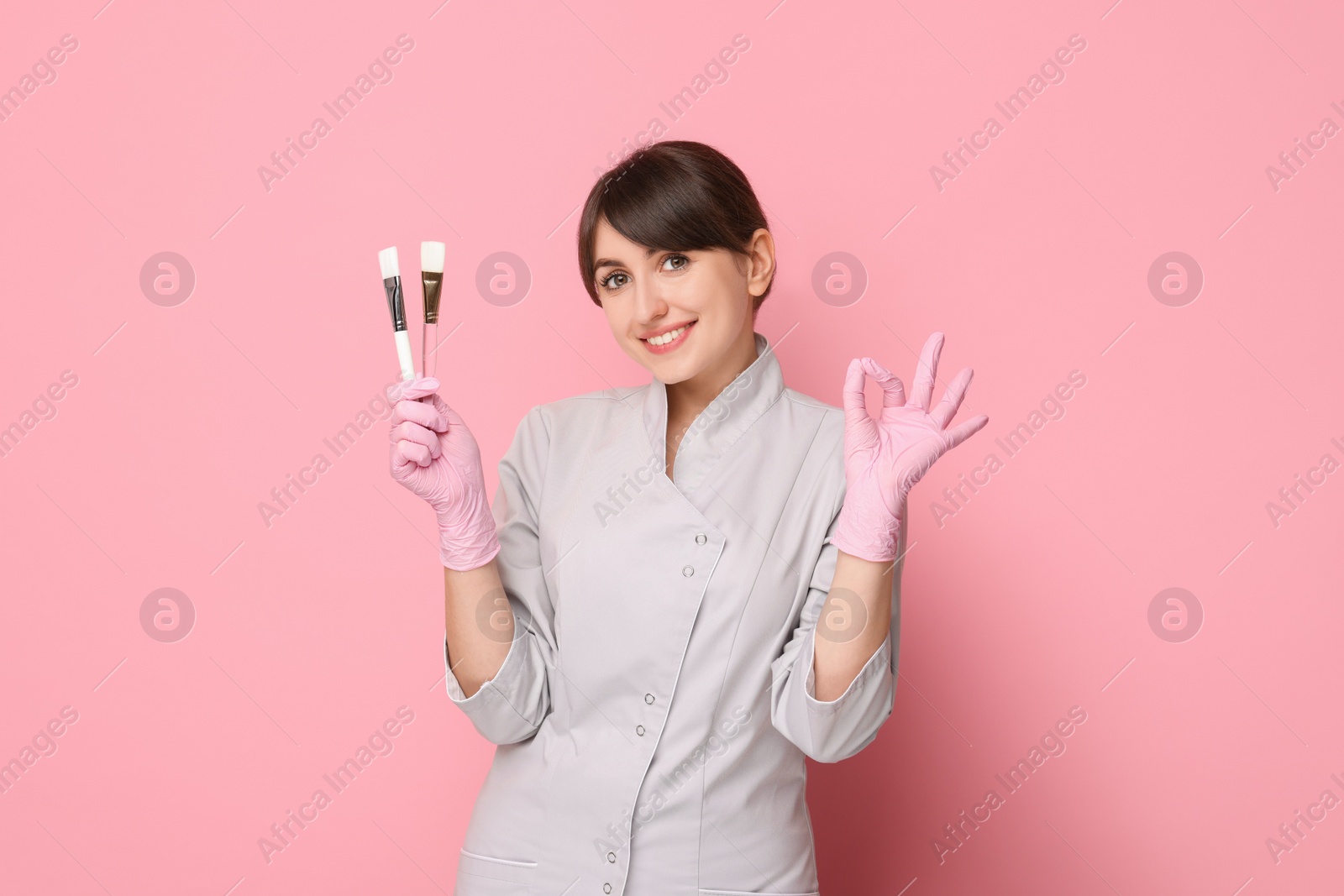 Photo of Cosmetologist with cosmetic brushes showing OK gesture on pink background