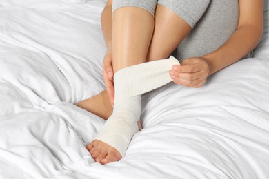 Photo of Young woman applying medical bandage onto foot on bed, closeup