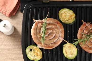 Photo of Electric grill with homemade sausages, zucchini and rosemary on wooden table, flat lay