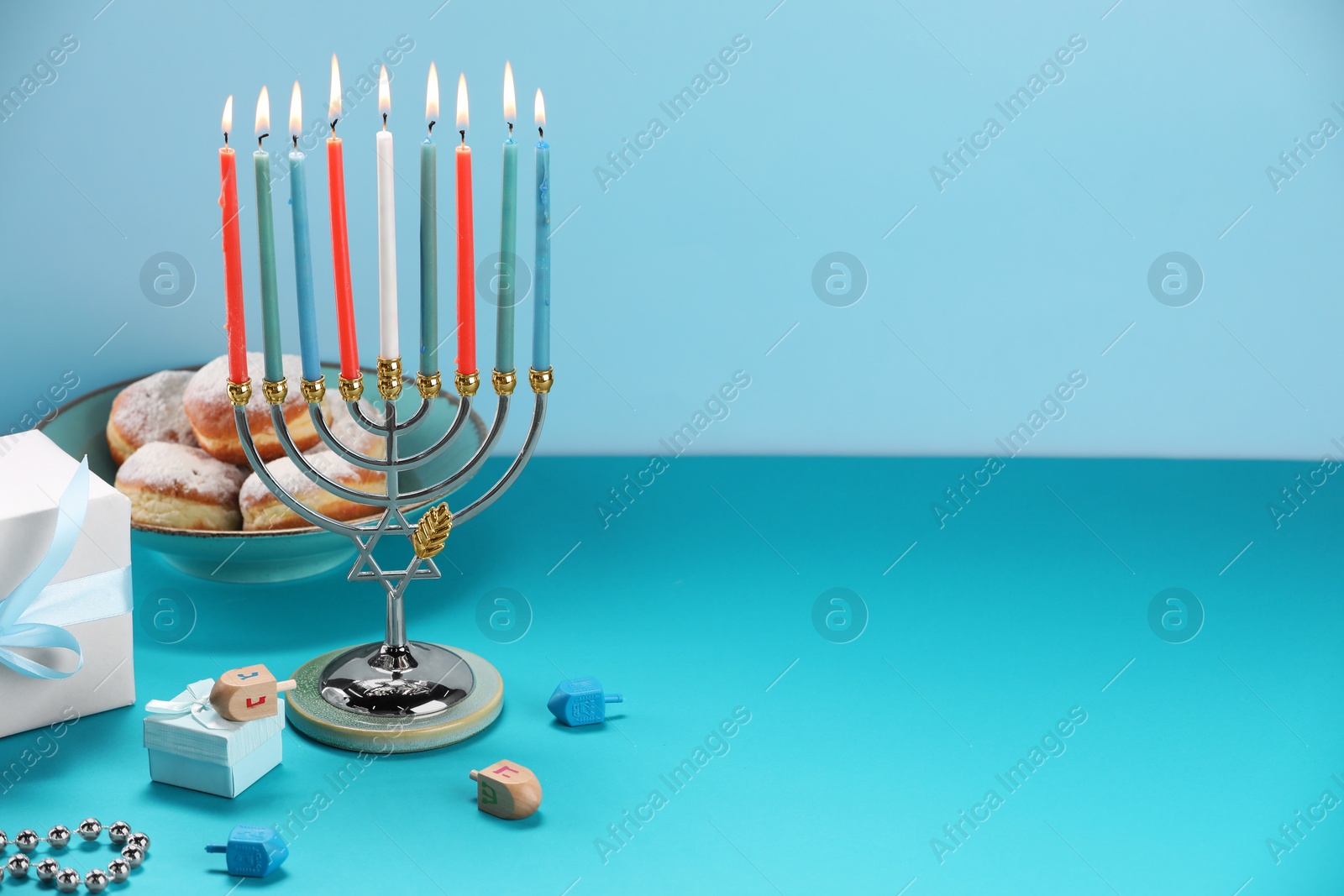 Photo of Hanukkah celebration. Menorah with burning candles, dreidels, donuts and gift boxes on table against light blue background. Space for text