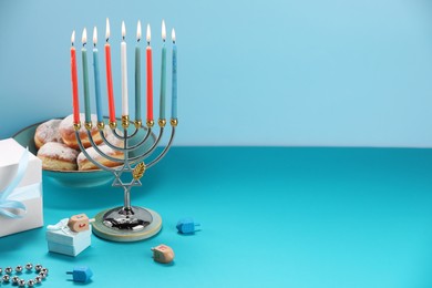 Photo of Hanukkah celebration. Menorah with burning candles, dreidels, donuts and gift boxes on table against light blue background. Space for text