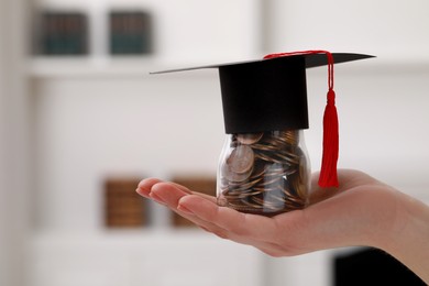 Woman holding glass jar of coins and graduation cap indoors, closeup with space for text. Scholarship concept