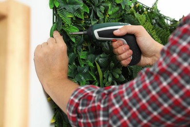 Photo of Man with screwdriver installing green artificial plant panel on white wall, closeup