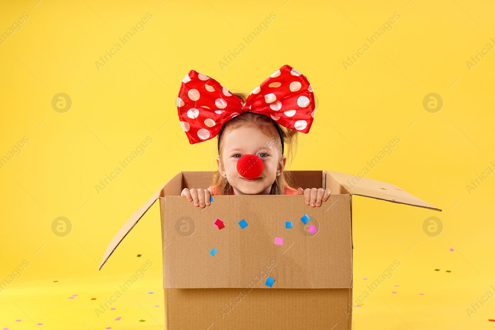 Photo of Little girl with large bow and clown nose in cardboard box on yellow background. April fool's day