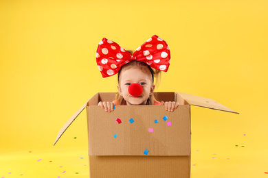 Photo of Little girl with large bow and clown nose in cardboard box on yellow background. April fool's day