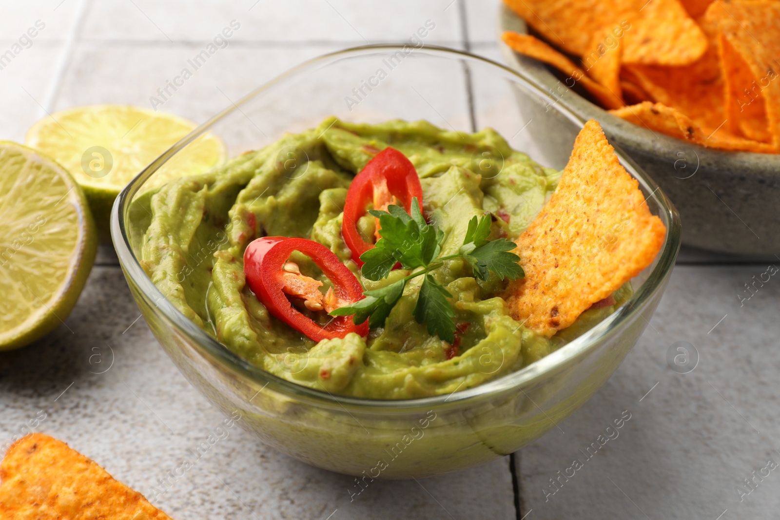Photo of Bowl of delicious guacamole with chili pepper, nachos chips and lime on white tiled table, closeup
