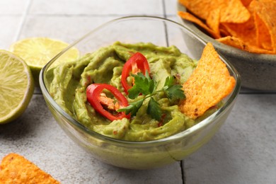 Bowl of delicious guacamole with chili pepper, nachos chips and lime on white tiled table, closeup