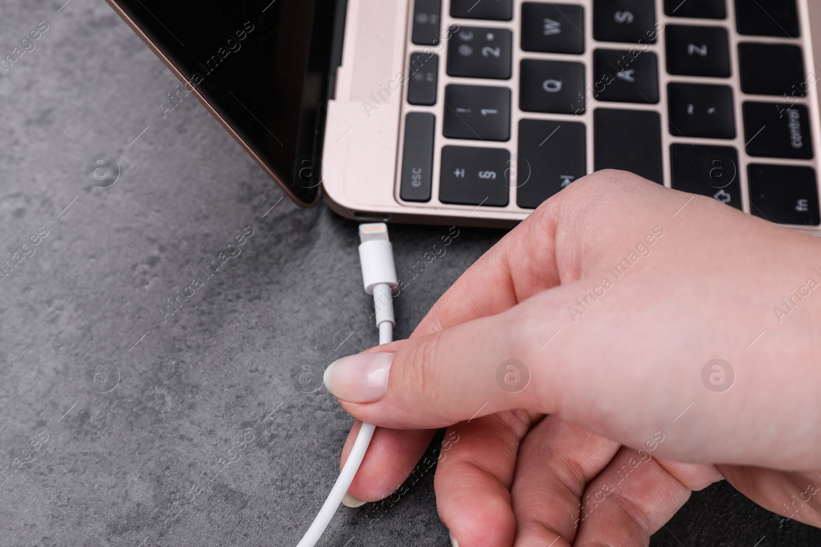 Photo of Woman plugging USB cable with lightning connector into laptop port on dark table, closeup