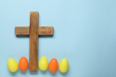 Wooden cross and painted Easter eggs on light blue background, flat lay. Space for text