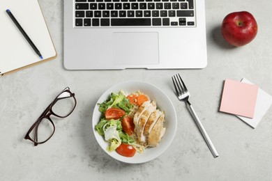 Photo of Bowl with tasty food, laptop, glasses, fork, apple and notebook on light grey table, flat lay. Business lunch