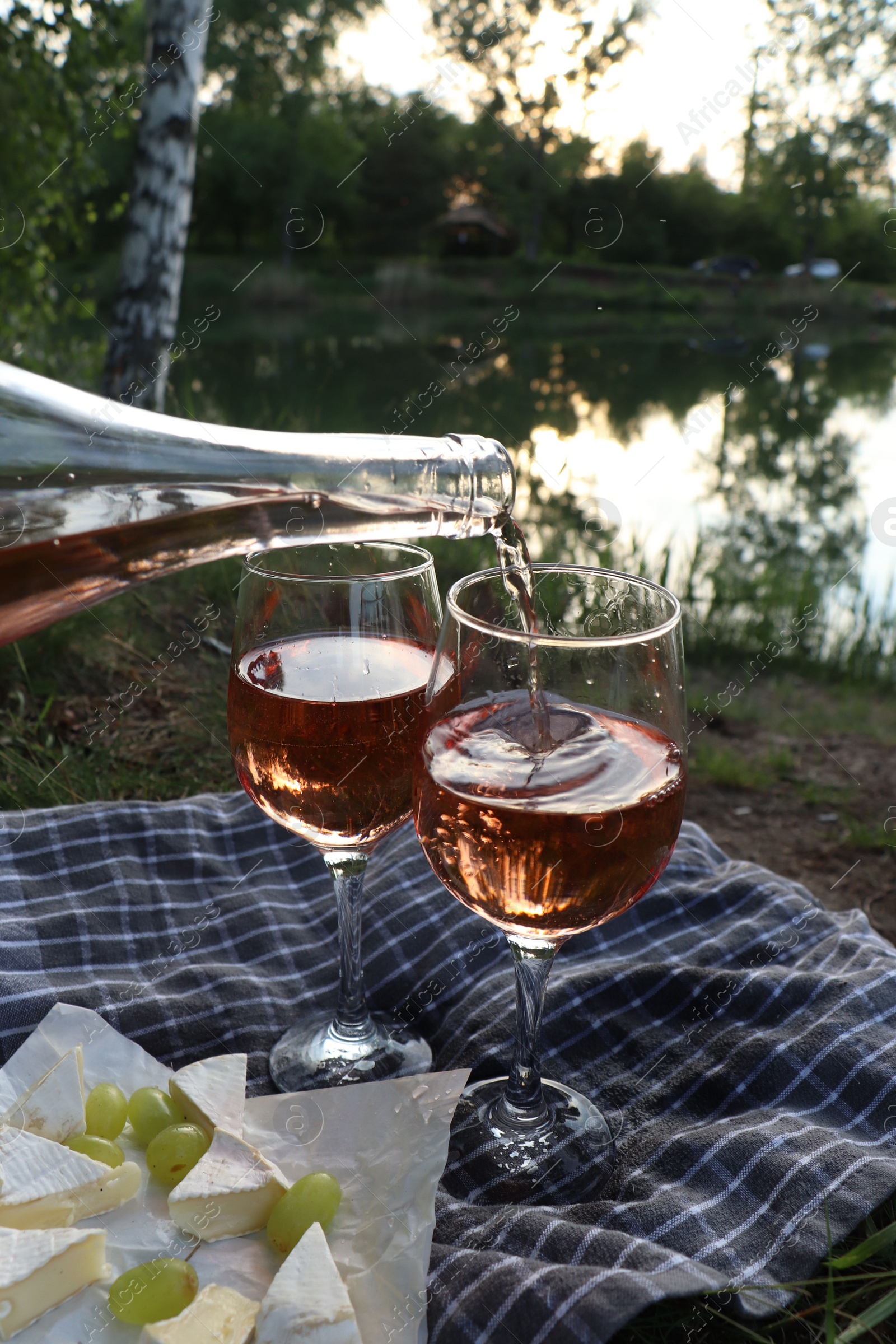 Photo of Pouring delicious rose wine into glass on picnic blanket near lake, closeup