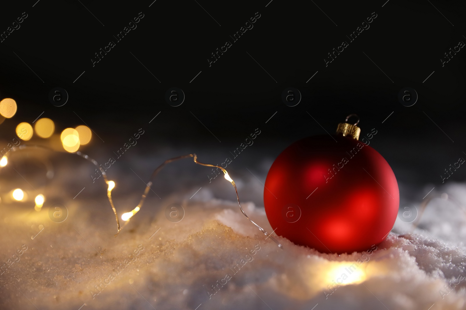 Photo of Christmas ball and fairy lights on white snow outdoors. Space for text