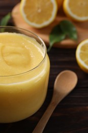 Photo of Delicious lemon curd in glass jar, fresh citrus fruit and spoon on wooden table, closeup