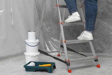 Photo of Woman climbing metallic folding ladder, tray with roller and buckets of paint indoors, closeup