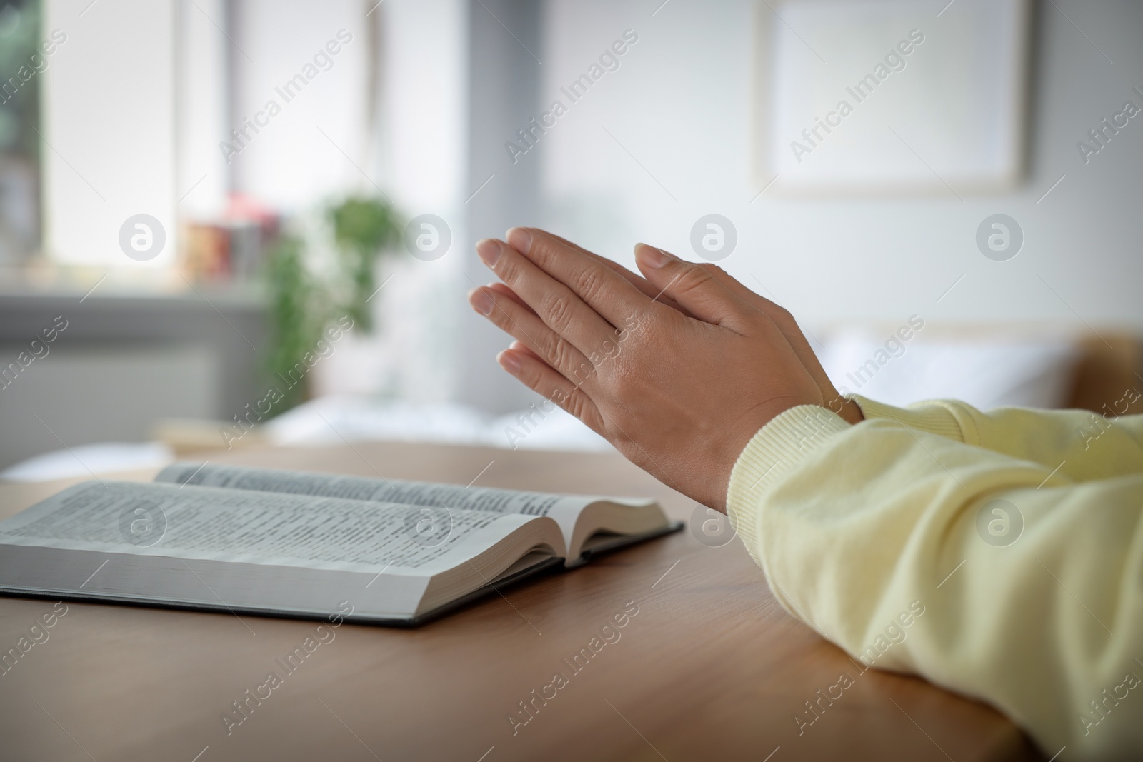 Photo of Woman holding hands clasped while praying at wooden table with Bible indoors, closeup. Space for text