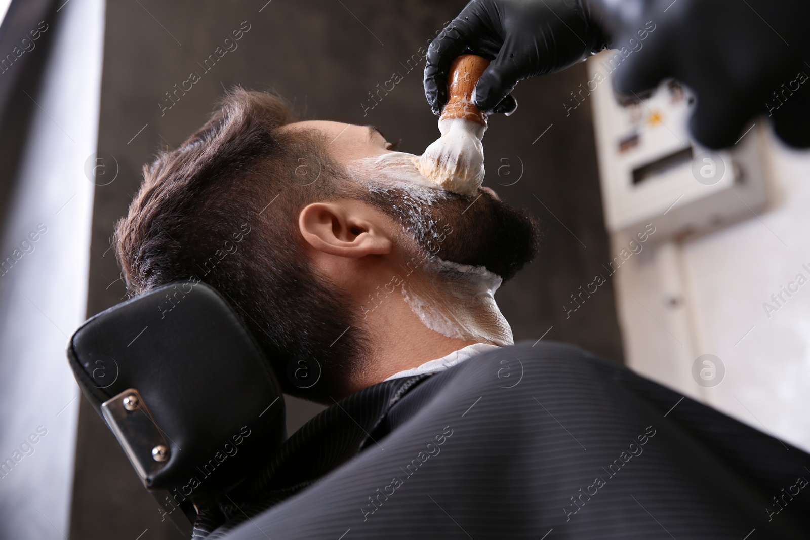 Photo of Professional hairdresser applying shaving foam onto client's skin in barbershop