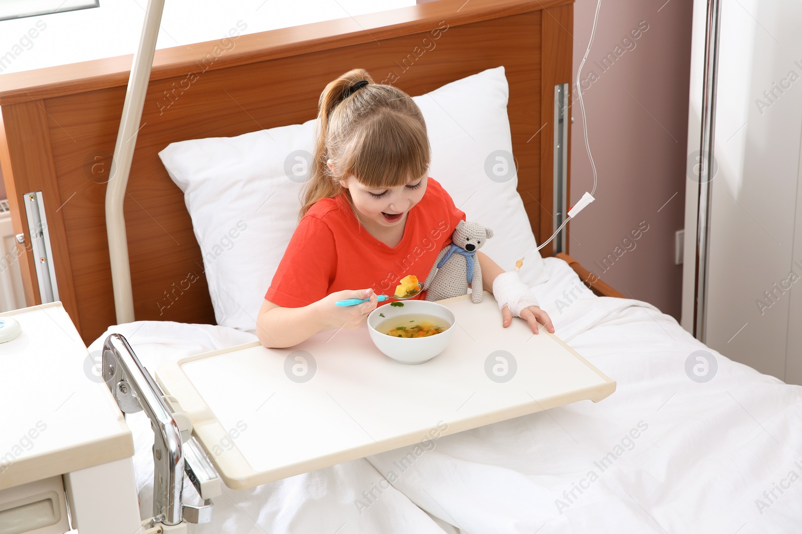 Photo of Little child with intravenous drip eating soup in hospital bed
