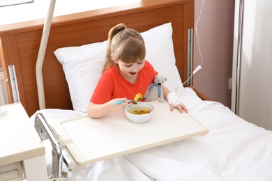 Photo of Little child with intravenous drip eating soup in hospital bed