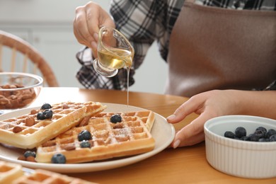 Photo of Woman pouring honey onto delicious Belgian waffles  at wooden table in kitchen, closeup