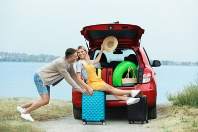 Photo of Couple having fun near car trunk with suitcases on riverside