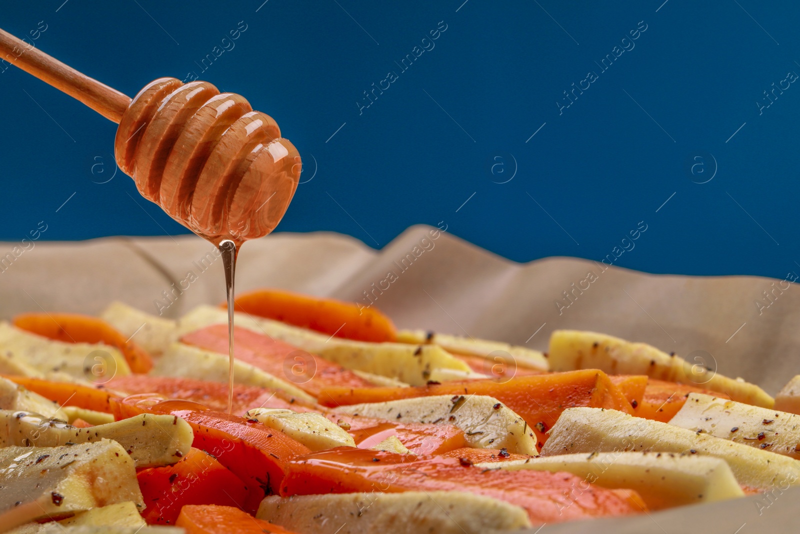 Photo of Pouring honey onto slices of parsnip and carrot against blue background, closeup