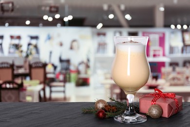 Image of Glass of delicious eggnog, Christmas ornaments, fir tree branches and gift box on dark table in bar, space for text