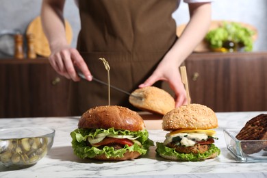 Photo of Woman cutting bun at white marble table, focus on delicious vegetarian burgers