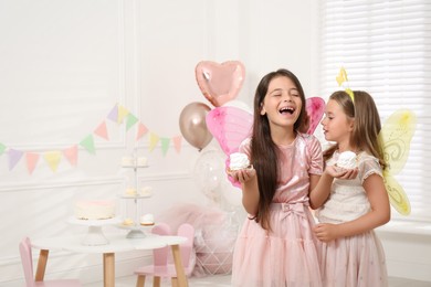 Photo of Cute little girls wearing fairy costumes with delicious cupcakes in decorated room. Space for text