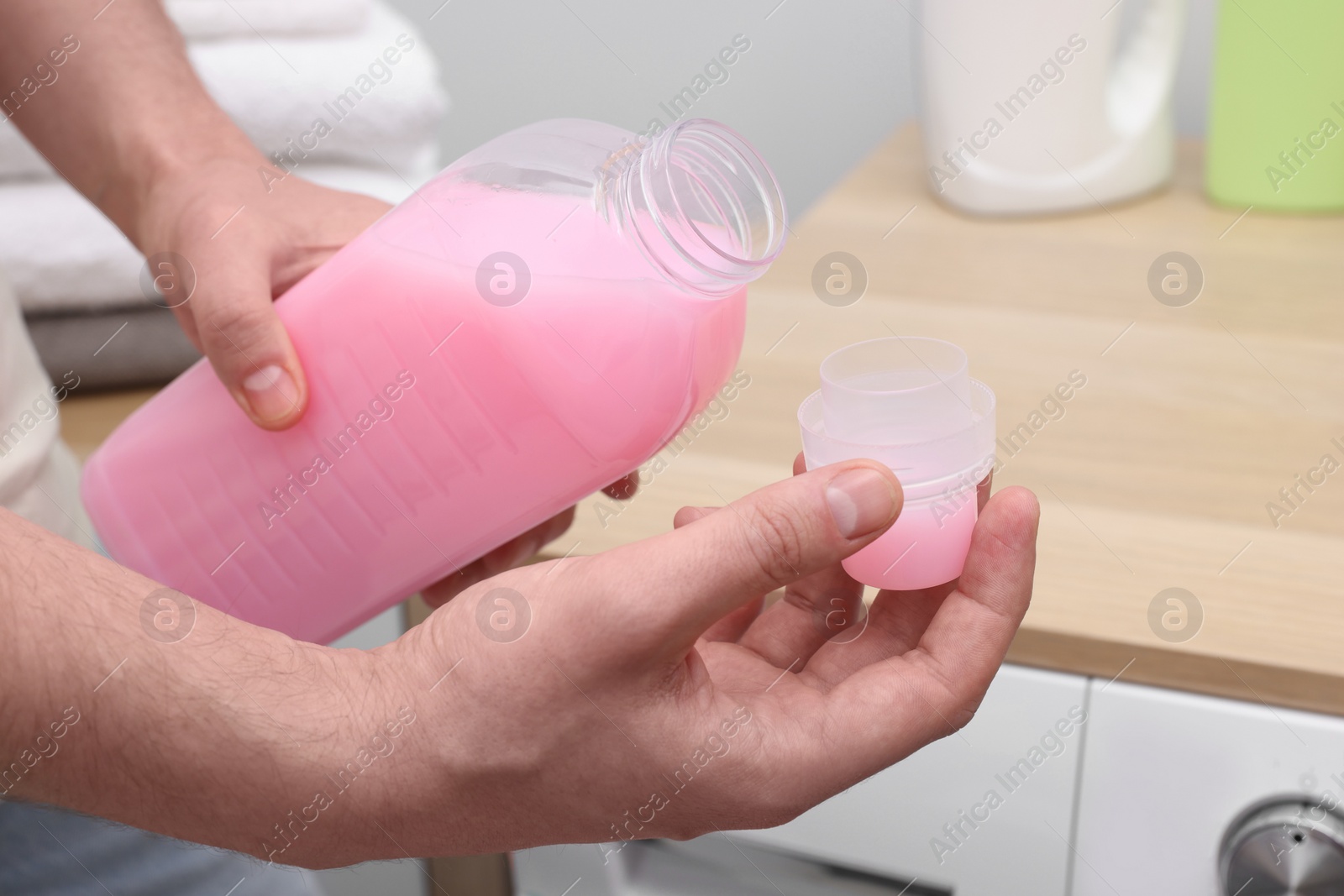 Photo of Man pouring fabric softener from bottle into cap near washing machine indoors, closeup