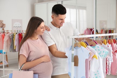 Happy pregnant woman with her husband choosing baby clothes in store. Shopping concept